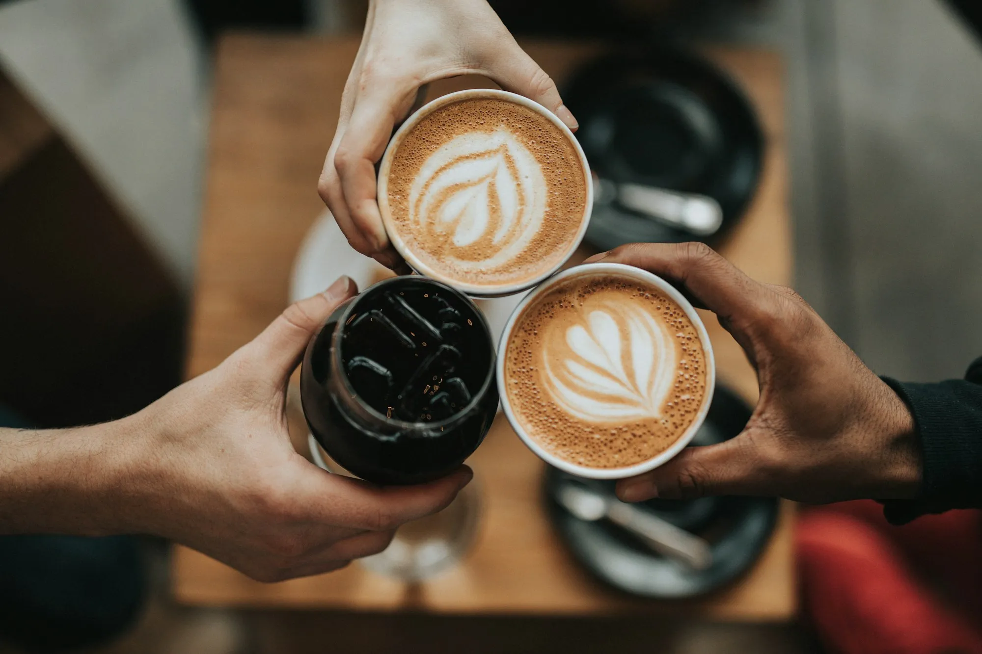 Zwei Hände stoßen mit Kaffeetassen mit Latte-Art-Muster über einem Holztisch an.